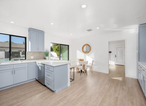 Modern kitchen interior with light blue cabinets, wooden flooring, and natural light streaming through large windows.