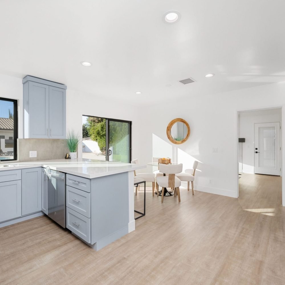 Modern kitchen interior with light blue cabinets, wooden flooring, and natural light streaming through large windows.